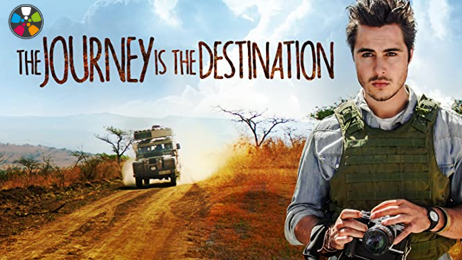 A white man wearing a bullet proof vest is on the front right hand side of the frame, a dirt road and blue sky behind him. Text reads: The Journey is the Destination