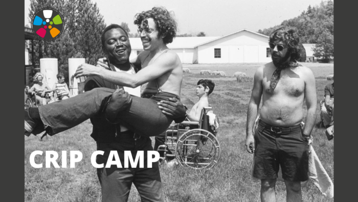 Black and white photograph from Camp Jened. In the photo, a Black camp counselor joyfully holds a white counselor in his arms. Both individuals are smiling. A shirtless white man looks on. A shirtless white camper in a wheelchair is in the background. They are all outdoors. The multicolored Journeys in Film circular logo is in the upper left-hand corner.