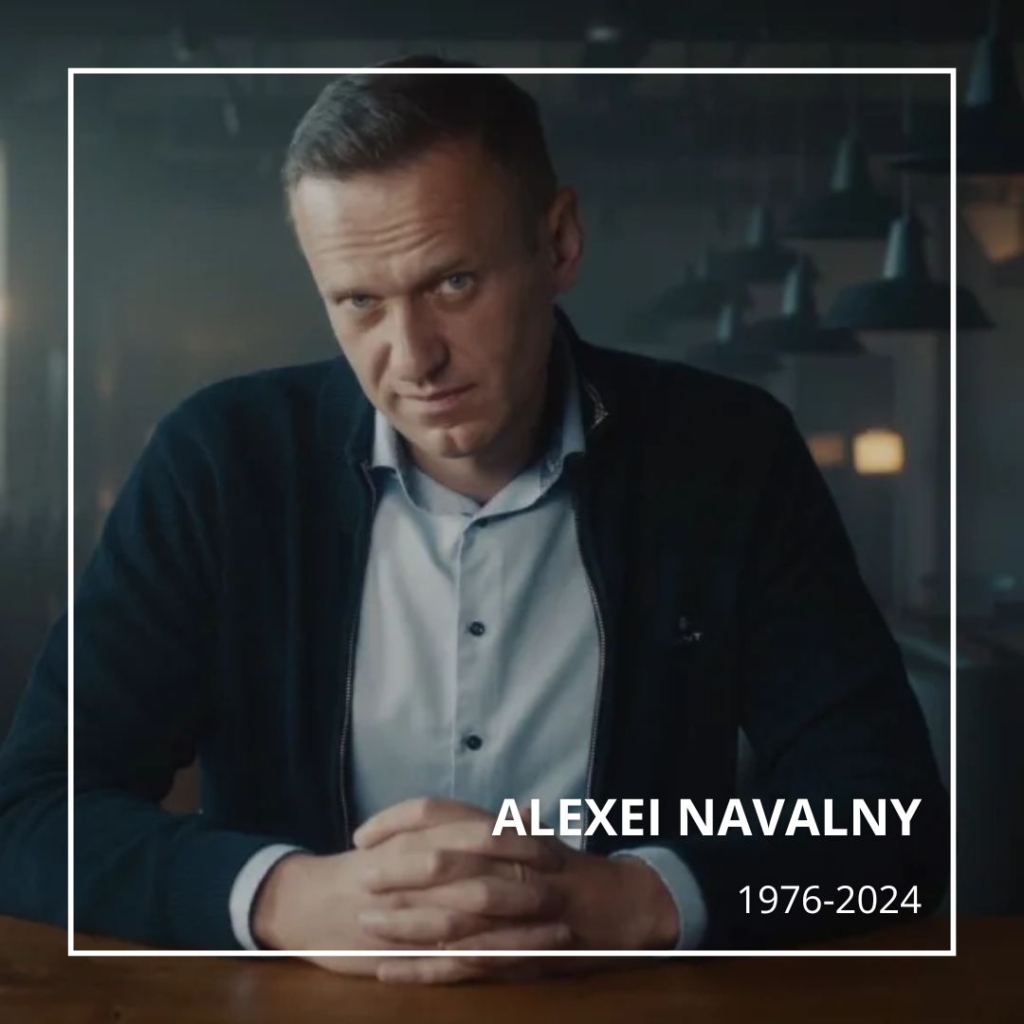 Still photograph from the 2022 documentary "Navalny", showing Alexei Navalny, a slim, middle-aged Russian white man with sandy brown hair, seated at a table. He wears a blue cardigan over a button up shirt. His hands are crossed in front of him. His head is tilted and he looks directly at the camera. Text says: "Alexei Navalny 1976-2024".