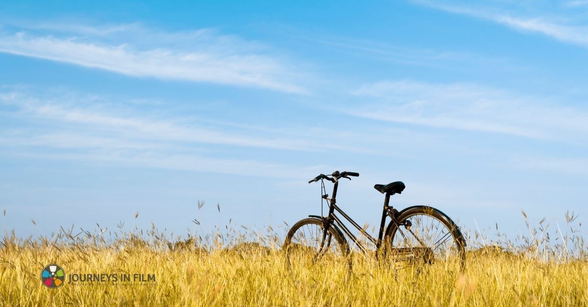 An old-fashion black bicycle in a dry field on a sunny day under a hazy blue sky. It makes you want to go on a bike ride! The Journeys in Film logo is in the bottom left corner.