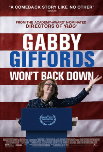 Film poster for Gabby Giffords Won't Back Down. Giffords, a middle aged white woman in a blue suit and glasses, speaks joyfully, her arm outstretched, at a blue podium in front of the red and white stripes of a huge American flag.