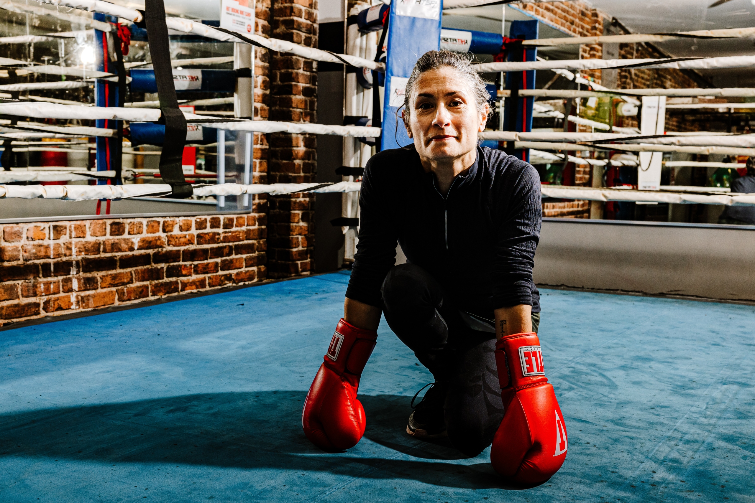 Middle age woman crouches in the center of the boxing ring. She wears red boxing gloves with the globes touching down on the mat.