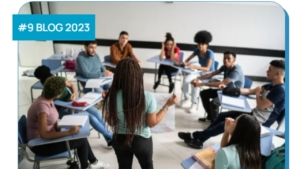 A photo sows a young woman in box braids from the back. She is holding up a large piece of paper, and addressing a diverse group of engaged students who sit at desks in a circle around her.