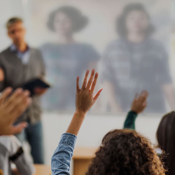 Class with hands raised while teacher is showing a film in the background.