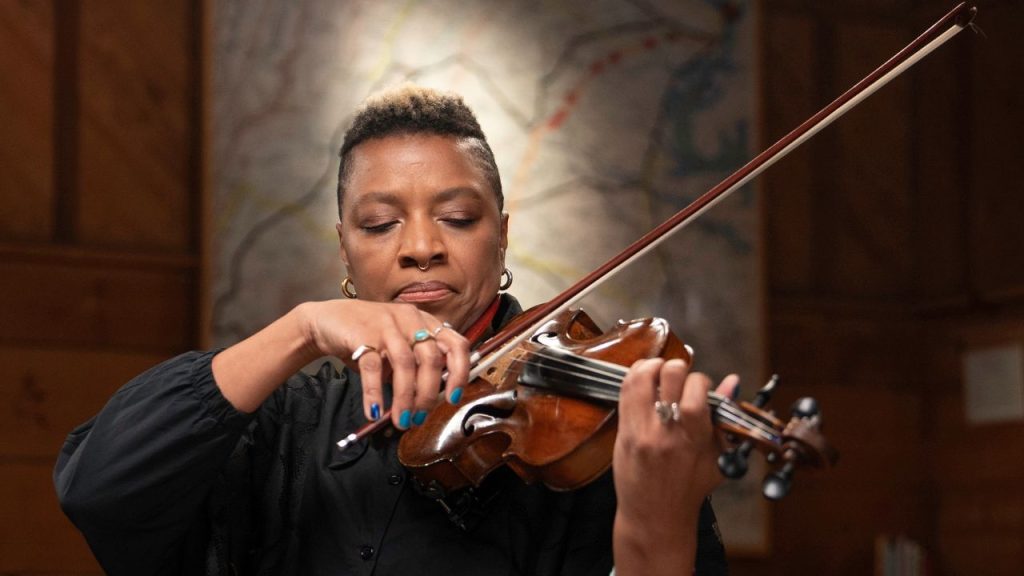Headshot of Black American Musician Mazz Swift, who uses they/them pronouns. They stand under a spotlight, playing a violin, with an expression of passionate concentration.