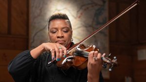 Headshot of Black American Musician Mazz Swift, who uses they/them pronouns. They stand under a spotlight, playing a violin, with an expression of passionate concentration.